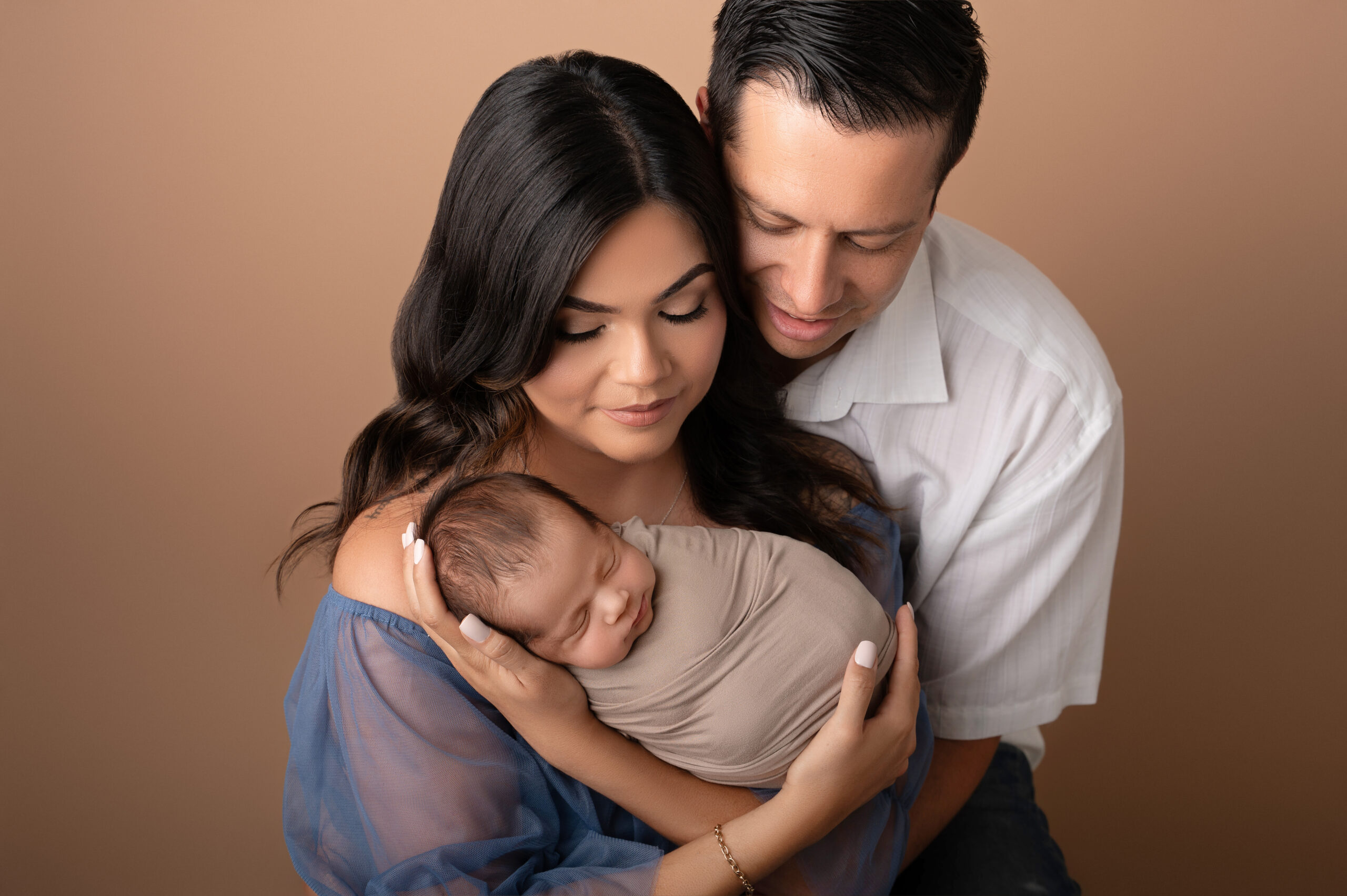 a woman and man lovingly cradle a sleeping newborn wrapped in a beige blanket, posing closely together against a plain tan background—an intimate moment beautifully captured by a sacramento baby portrait studio.