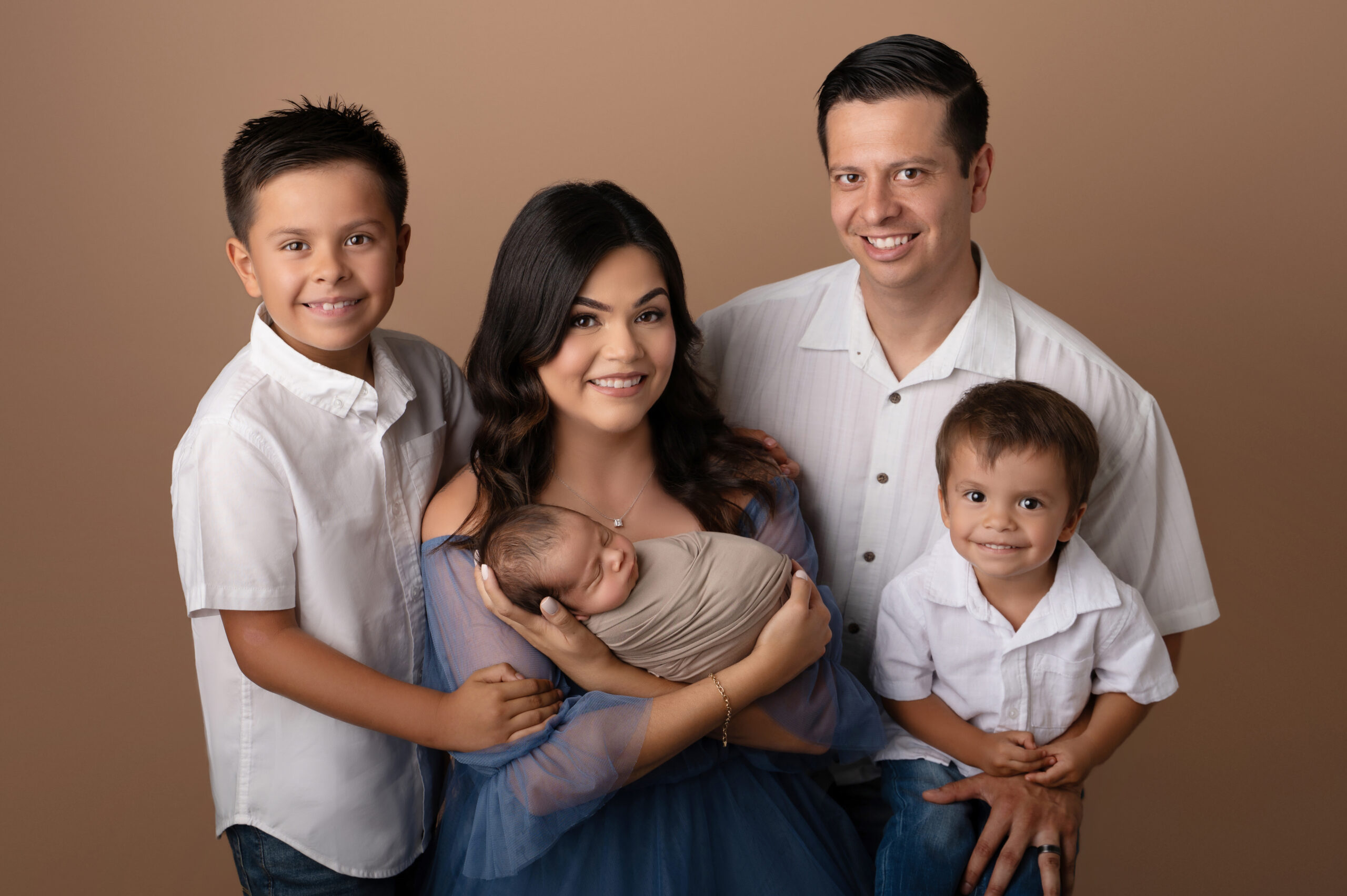 a family of five poses together. a mother in a blue dress holds a swaddled baby, capturing the joy of baby milestone photos sacramento families treasure. two young boys and a man in white shirts stand smiling around them in front of a tan background.