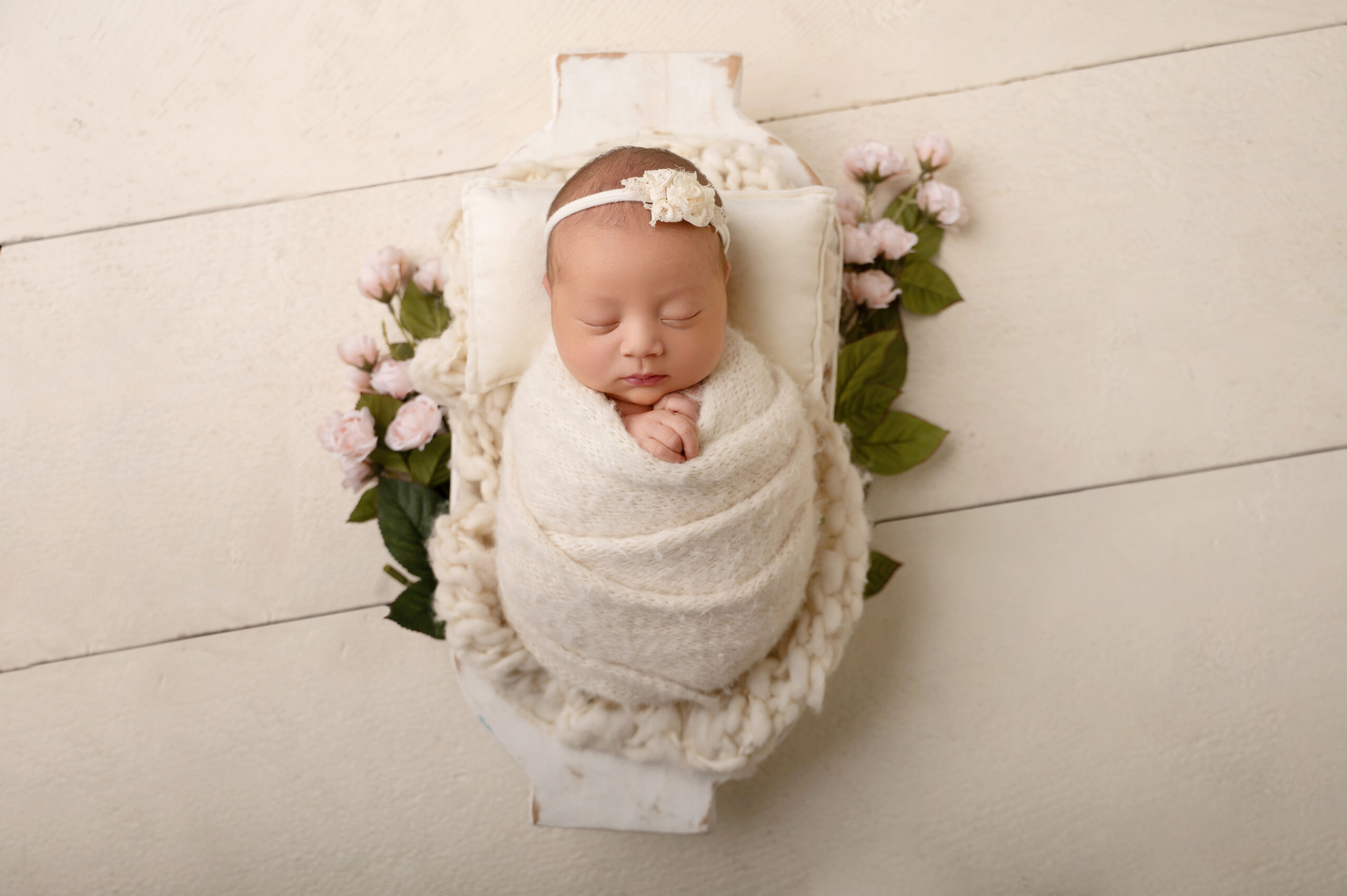 newborn baby wrapped in a cream blanket and headband, sleeping on a small white bed, surrounded by pink flowers and green leaves, on a light wooden floor—a perfect moment captured by an experienced sacramento baby photography studio.