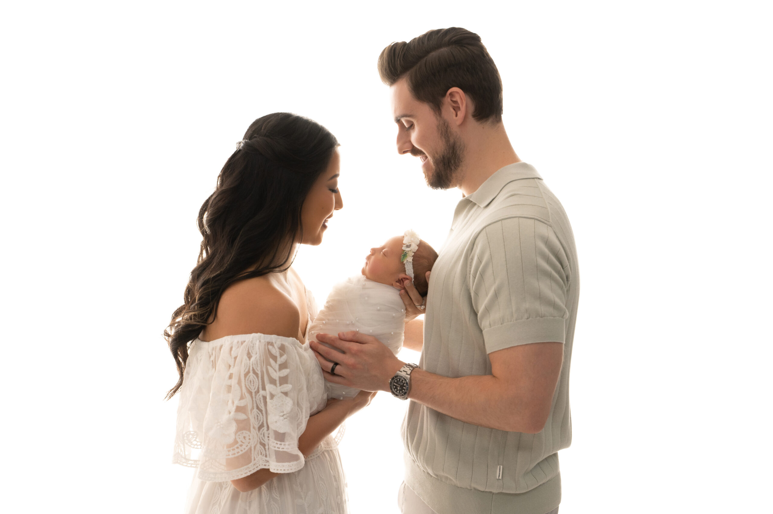 a woman and man smile while holding a swaddled newborn baby, facing each other against a white background—a heartwarming moment beautifully captured by sacramento baby photography.