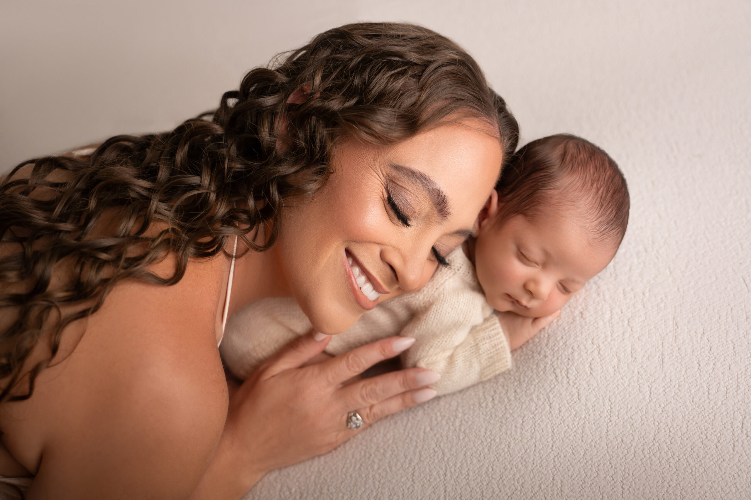 smiling woman with long curly hair gently embraces a sleeping newborn on a soft, light colored blanket. both have peaceful expressions and are lying close together—capturing the pure love and tenderness in this newborn photoshoot sacramento.